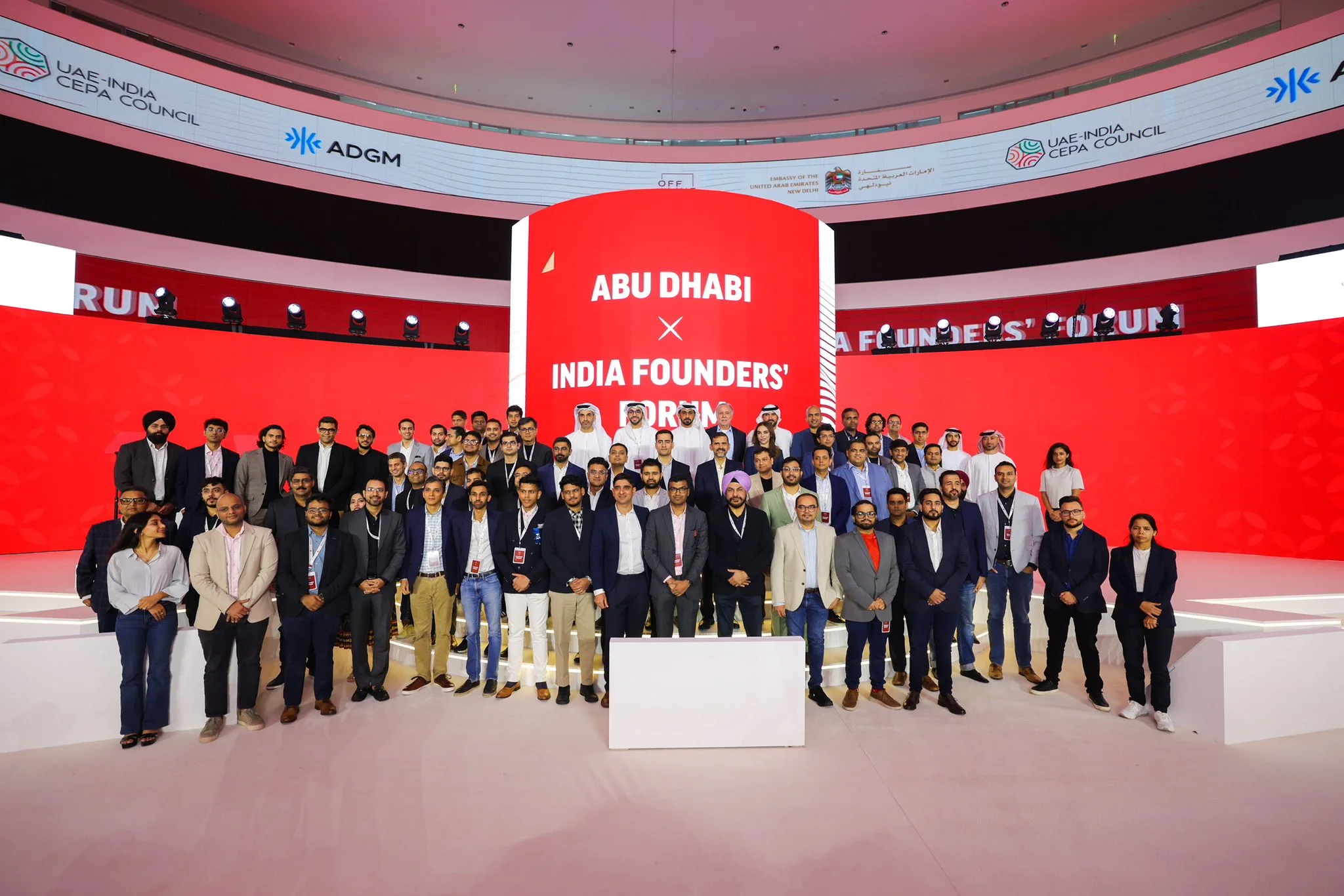 The Founders’ Forum stage: a curved red LED wall around a red cylinder reading “Abu Dhabi × India Founders’ Forum”, with a ring of partner logos above and a large group of delegates in front.