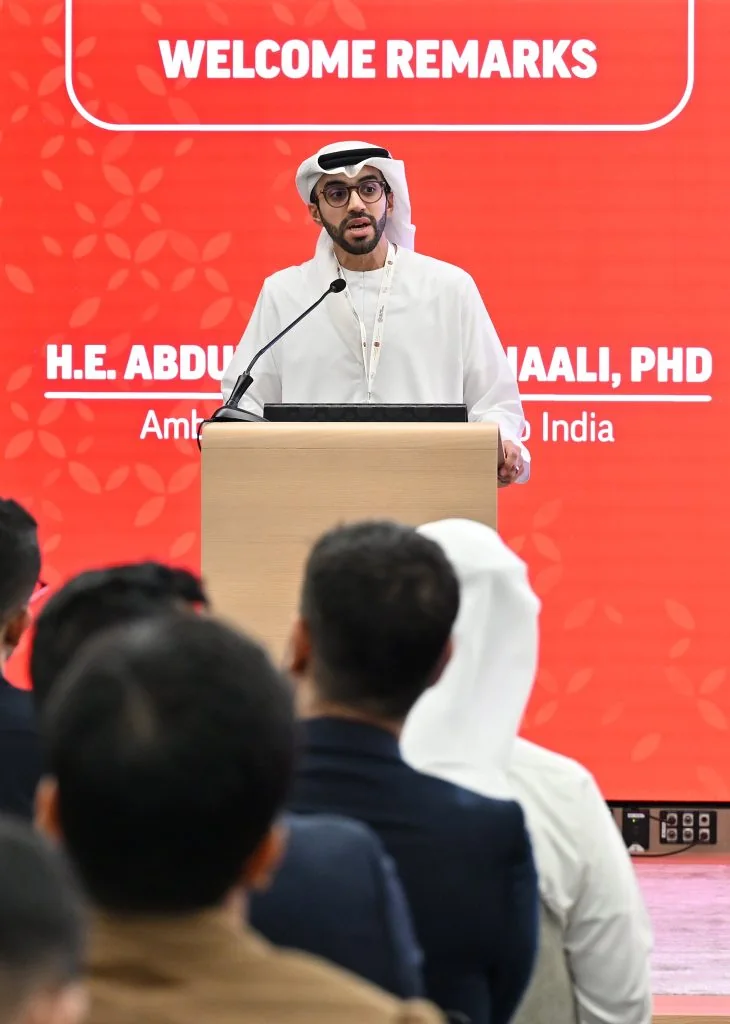 A speaker at a lectern under a red LED name card with a “Welcome Remarks” label, seen over the heads of the audience.